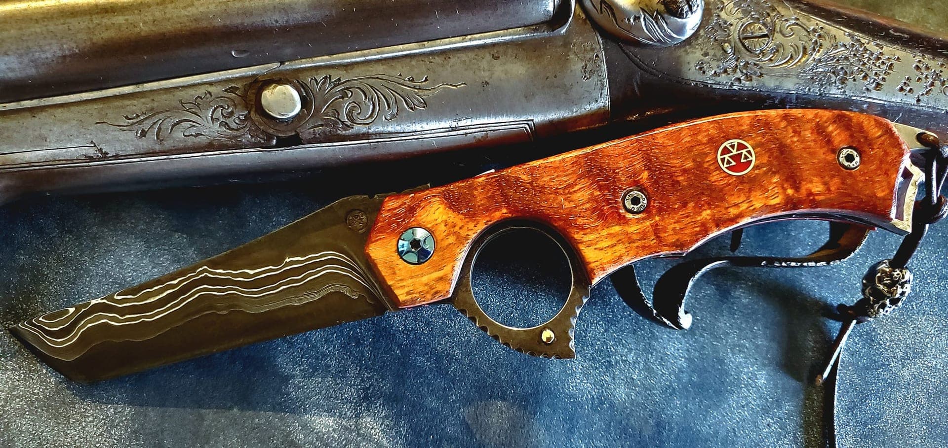 L'Archétype, couteau damas à manche en loupe de bois, posé près d'un fusil ancien gravé.
