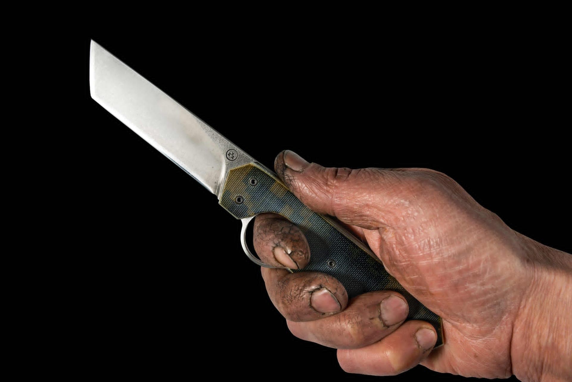 A worker's hand, marked and calloused, holding the Résiliant Tanto open against a black background, index finger through the reverse-opening ring.