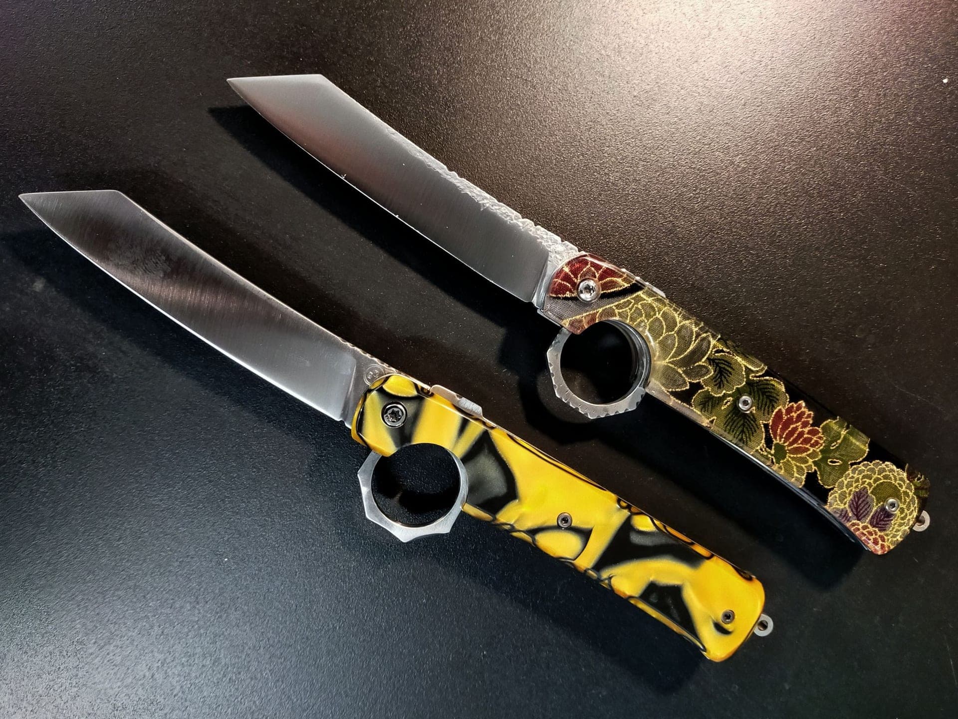 Deux Shotokan ouverts côte à côte sur fond sombre, lames claires à dos droit et anneaux d'ouverture au talon, l'un à manche jaune et noir, l'autre à manche sombre à motif floral.