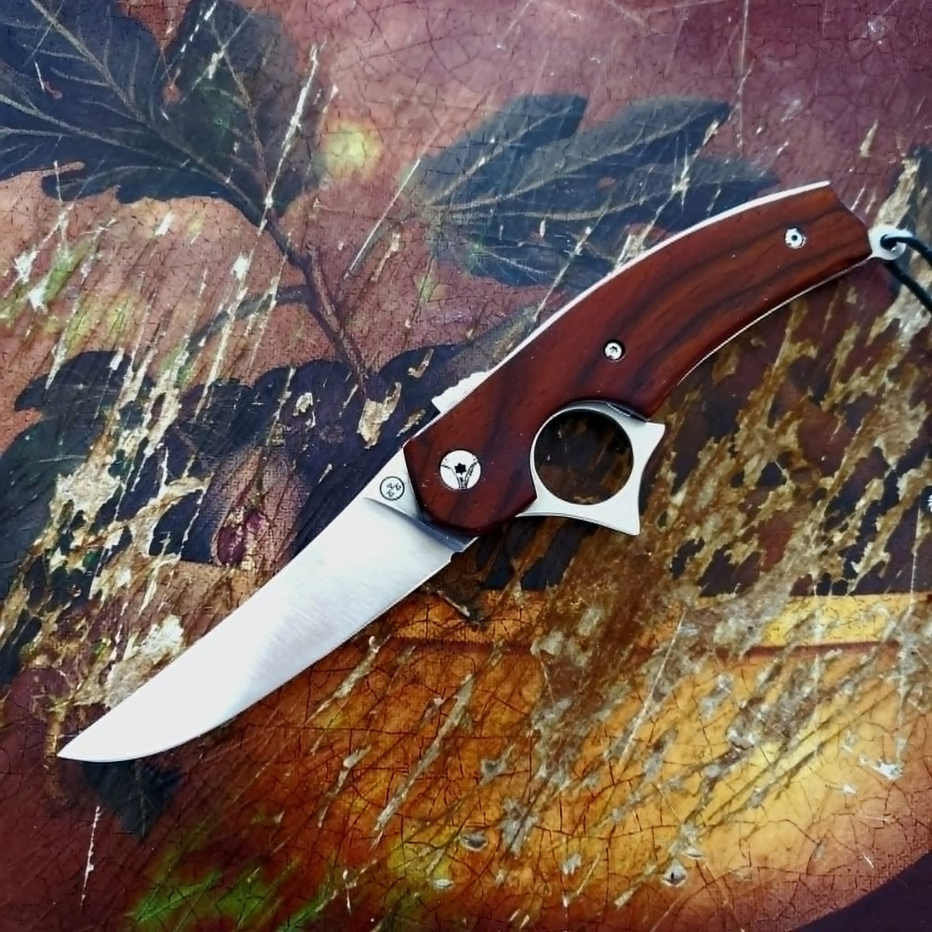 Le Tracker, couteau à ouverture inversée à lame inox 14C28N polie et manche en bois veiné rougeâtre, ouvert sur un fond peint patiné.