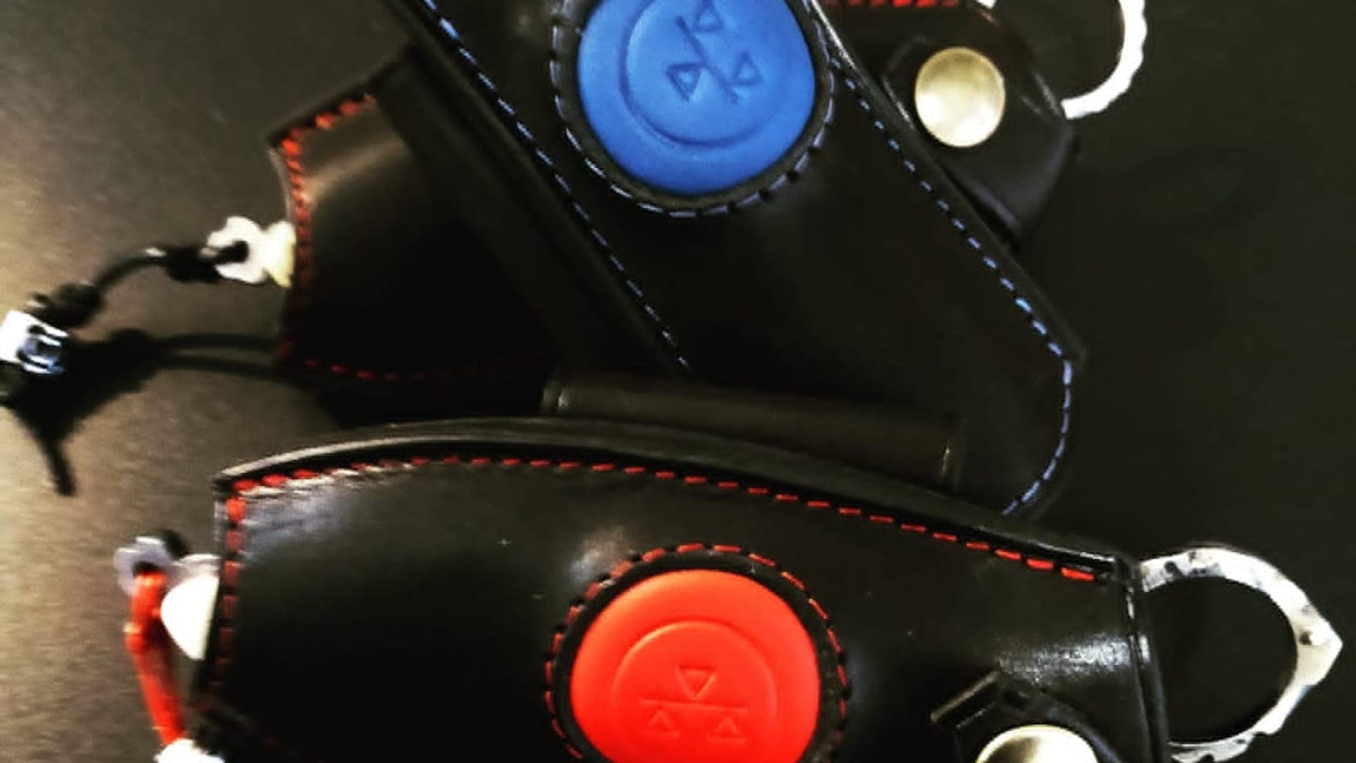Deux étuis du Résiliant en cuir noir cousu main, vus de près, l'un à pastille bleue, l'autre à pastille rouge au logo, surpiqûres assorties et bouton-pression.