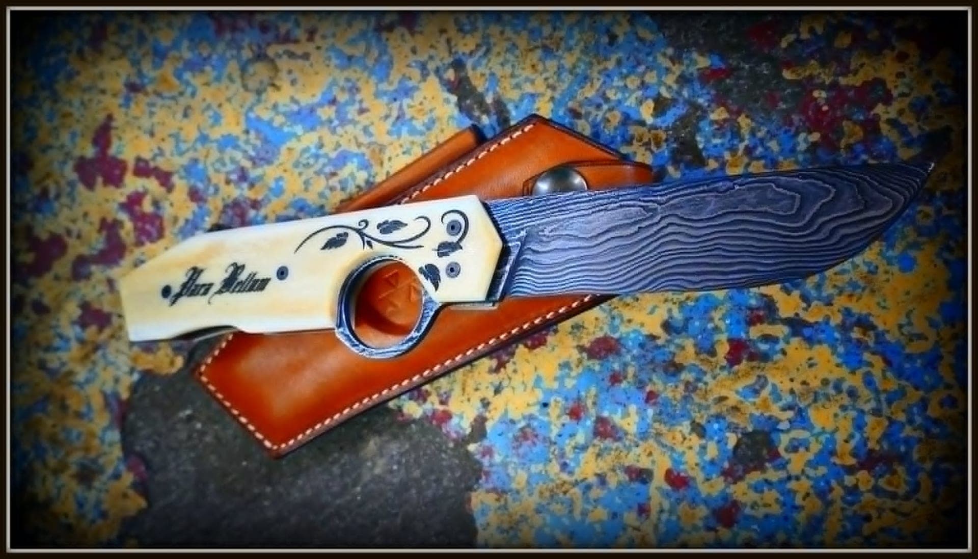 A Résiliant 2.0 with a damascus blade and a pale handle engraved with its owner's name, resting on its hand-stitched tan leather sheath.